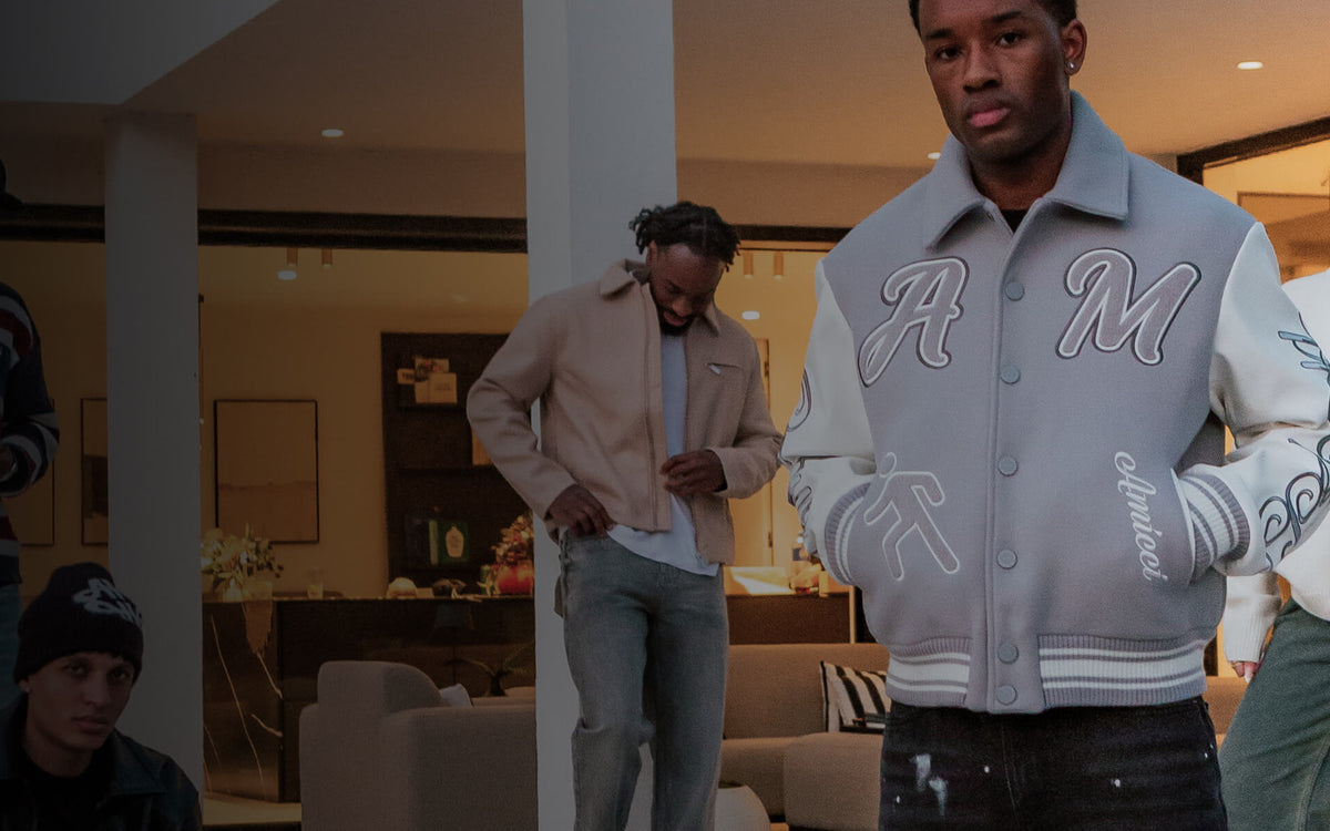 A group of people standing in a modern indoor living space, with the central focus on a man wearing a light gray varsity-style jacket with large white 'AM' lettering on the front and sleeves, alongside other casually dressed individuals.